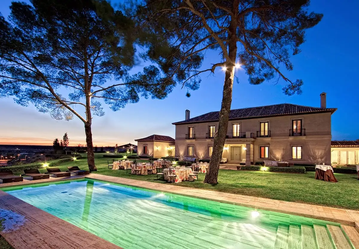 La casa de dos plantas al anochecer, con la piscina iluminada, dos pinos y mesas puestas sobre el césped
