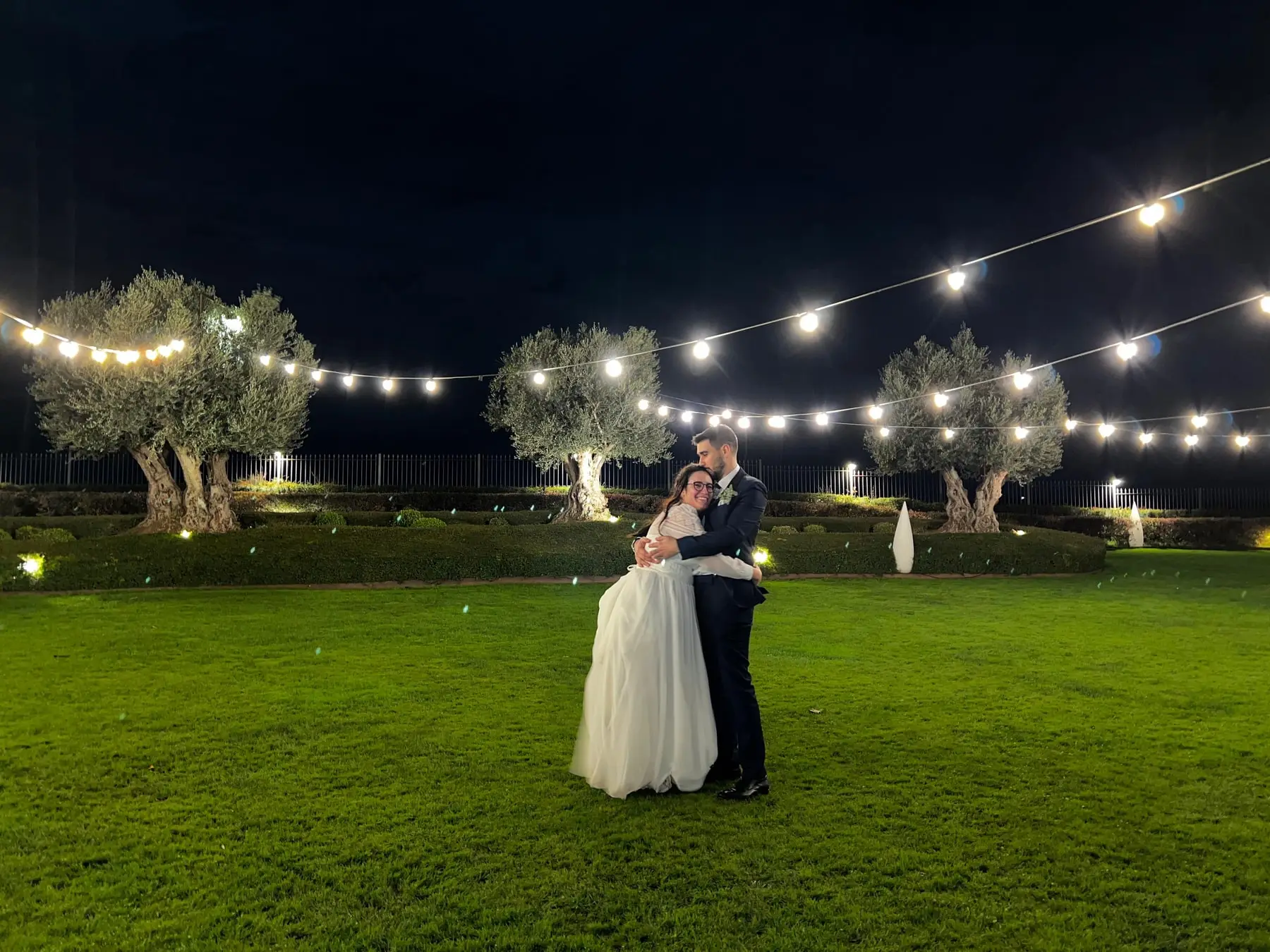 Los novios abrazados sobre el césped de noche, bajo guirnaldas de luces y olivos